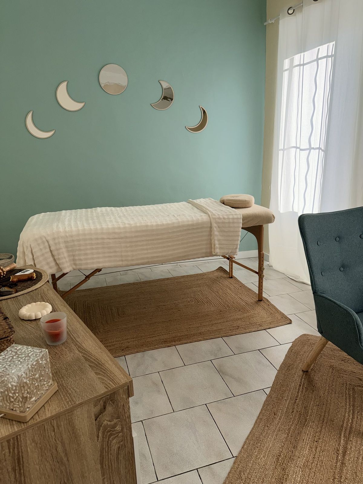 Table de massage avec linge écru, tapis de jute et phases de lune en miroir sur le mur vert d'eau du cabinet